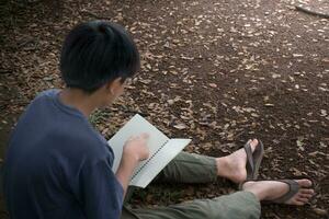 asiatico giovane ragazzo seduta lettura libro a terra nel giardino, bambini lettura libro e imparando, ragazzo con prenotare, concetto di formazione scolastica e naturale, terra e tramonto contesto, sostenibilità. foto