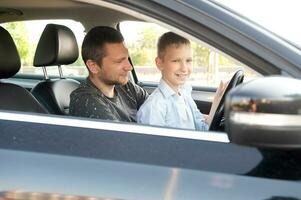 papà e figlio nel il davanti posto a sedere di un' auto guida un' nero macchina. il ragazzo detiene il timone ruota. foto