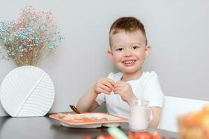 ridendo ragazzo mangia delizioso pasta a il tavolo nel il cucina foto