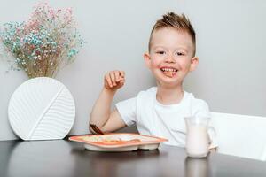 ridendo ragazzo mangia delizioso pasta a il tavolo nel il cucina foto