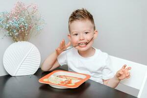 contento bambino mangiare delizioso pasta a il tavolo nel il cucina foto