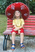 poco carino ragazza indossare un' grande sombrero è seduta su un' rosso panchina nel un' pubblico parco foto