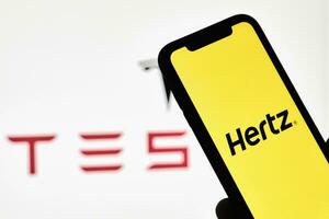 un' persona detiene smartphone con hertz marchio al di sopra di grande tesla logo foto