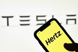 un' persona detiene smartphone con hertz marchio al di sopra di grande tesla logo foto