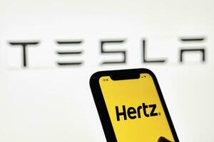 un' persona detiene smartphone con hertz marchio al di sopra di grande tesla logo foto