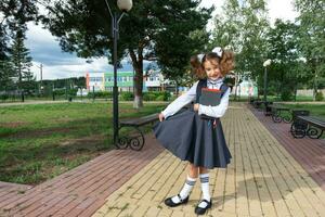 ragazza con zaino, scuola uniforme con bianca fiocchi e pila di libri vicino scuola. indietro per scuola, contento allievo, pesante libri di testo. formazione scolastica, primario scuola classi, settembre 1 foto