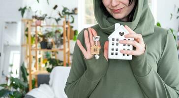 un' contento donna nel sua Casa detiene nel sua mani un' miniatura figura di un' Casa e chiave nel il interno. sognare Casa progetto, vero tenuta Acquista, assicurazione, mutuo, affitto, prenotazione foto