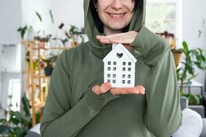 un' contento donna nel sua Casa detiene nel sua mani un' miniatura figura di un' Casa e chiave nel il interno. sognare Casa progetto, vero tenuta Acquista, assicurazione, mutuo, affitto, prenotazione foto