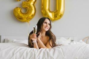bellissimo donna svegliato su nel letto nel il mattina e celebra sua anni trenta con un' bicchiere di Champagne nel un' camicia da notte foto