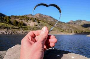 un' persona Tenere un' cuore sagomato oggetto nel davanti di un' lago foto