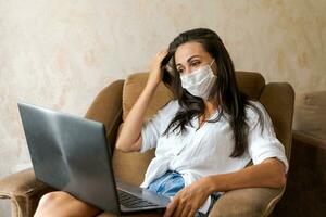 carino donna a casa nel sedia con il computer portatile indossare protettivo maschera su sua viso foto