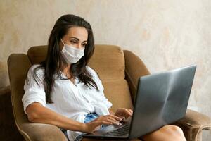 carino donna a casa nel sedia con il computer portatile indossare protettivo maschera su sua viso foto