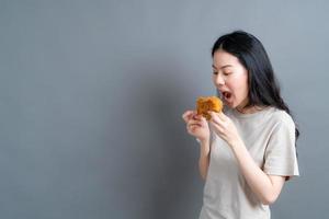 giovane donna asiatica che indossa t-shirt con faccia felice e si diverte a mangiare pollo fritto su sfondo grigio foto