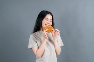 giovane donna asiatica che indossa t-shirt con faccia felice e si diverte a mangiare pollo fritto su sfondo grigio foto