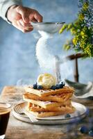 cialde sormontato con mirtilli, ghiaccio crema e miele in piedi su un' rustico Marrone tavolo. blu sfondo. mano spolverare zucchero polvere. foto
