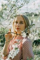 bionda ragazza su un' primavera camminare nel il giardino con ciliegia fiori. femmina ritratto, avvicinamento. un' ragazza nel un' rosa polka punto vestire. foto