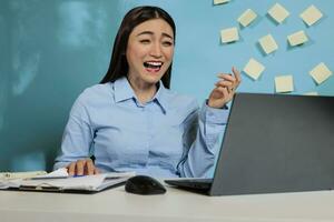 ottimista asiatico donna avendo un' divertente video chiamata conversazione seduta a ufficio scrivania. femmina azienda dipendente ridendo con eccitazione mentre parlando per collega a distanza. foto