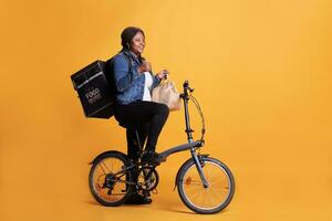 pizzeria dipendente Tenere carta Borsa pieno con porta via cibo ordine consegna per cliente durante pranzo volta, Corriere fare pollici su gesto a telecamera. fattorina utilizzando bicicletta come mezzi di trasporto foto