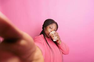 gioioso africano americano modello Tenere tazza potabile caffè durante video chiamata incontro utilizzando Telefono mentre in piedi nel studio al di sopra di rosa sfondo. donna avendo positivo emozione, godendo tempo libero tempo foto
