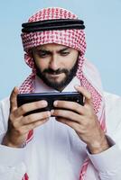 concentrato musulmano uomo indossare ghutra foulard giocando video gioco su smartphone. focalizzata arabo gamer vestito nel tradizionale Abiti godendo video gioco applicazione su mobile Telefono foto