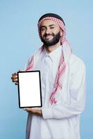 sorridente musulmano uomo mostrando digitale tavoletta con vuoto bianca schermo e guardare a telecamera con allegro espressione. contento arabo inserzionista Tenere vuoto touch screen ritratto foto