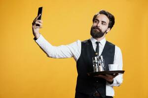 giovane Cameriere assunzione fotografie nel studio, utilizzando mobile Telefono ap per catturare sciocco immagini di lui con piatto nel mano. maggiordomo Lavorando a ristorante assunzione selfie contro giallo sfondo.