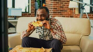 contento tipo mangiare fetta di Pizza e potabile birra a casa, godendo portare fuori cibo e Guardando televisione. giovane uomo servendo porta via cena e alcol, orologio film nel vivente camera. foto