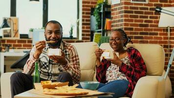 africano americano persone mangiare tagliatelle con bastoncini e abbuffata Guardando azione tv serie a casa. giovane partner sensazione rilassato mangiare asiatico porta via cibo, orologio film. tripode sparo. foto