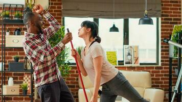 moderno sorridente coppia danza e pulizia appartamento, rastrellamento e passare l'aspirapolvere vivente camera di legno piani. giovane positivo partner avendo divertimento con danza si sposta, godendo lavori di casa. palmare sparo. foto