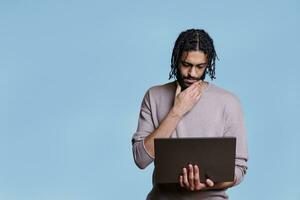 giovane riflessivo arabo uomo Tenere il computer portatile mentre analizzando Software codice e sfregamento mento. pensieroso persona pensiero nel dubbio mentre lettura attività commerciale rapporto su portatile computer foto