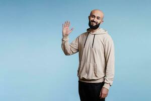 allegro arabo uomo sorridente e raccolta mano nel saluto mentre guardare a telecamera. giovane persona con contento facciale espressione accogliente e agitando Ciao con braccio studio ritratto foto