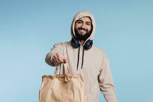 allegro sorridente arabo uomo nel cappuccio dando carta Borsa con prendere su cibo ritratto. ristorante consegna servizio Corriere Tenere pacchetto con porta via pranzo e guardare a telecamera foto