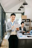 giovane attraente asiatico maschio ufficio lavoratore attività commerciale tute sorridente a telecamera con Lavorando bloc notes, tavoletta e il computer portatile documenti nel ufficio foto