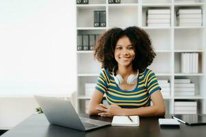 femmina studenti Nota a partire dal il libri a il africano ragazza biblioteca seduta a il scrivania utilizzando il computer portatile computer e tavoletta per ricerca un in linea informazioni. foto