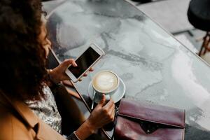 Visualizza a partire dal sopra. bellissimo giovane africano americano donna lettura testo Messaggio su mobile Telefono a caffè negozio. Tenere caldo bevanda. inverno stagione. foto