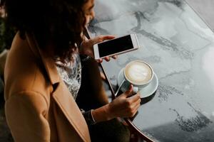 Visualizza a partire dal sopra. bellissimo giovane africano americano donna lettura testo Messaggio su mobile Telefono a caffè negozio. Tenere caldo bevanda. inverno stagione. foto