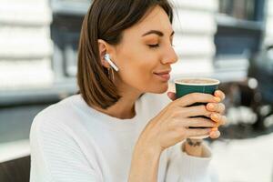 beato corto dai capelli donna godendo cappucino nel bar. indossare accogliente bianca maglione. ascoltando preferito musica di auricolari. foto