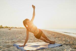 un' donna nel un' gli sport reggiseno e pantaloncini è fare yoga su il spiaggia foto