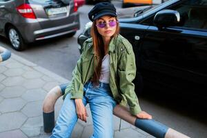 alla moda donna con lungo brunet capelli in posa all'aperto nel grande città vicino strada . indossare elegante verde giacca , nero berretto e viola occhiali da sole. foto