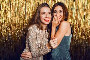 vicino su studio Immagine di Due sorprendente sexy festeggiare ragazze con rosso labbra, sorpresa viso, ridendo , in posa su d'oro scintillante sfondo. nuovo anno festa umore. foto
