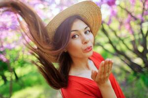 spensierato ragazza nel elegante cannuccia cappello e corallo vestito invio bacio per telecamera. rosa fioritura alberi su sfondo. indossare cannuccia cappello e corallo vestire. foto