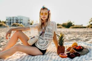 estate immagine di bello biondo donna Tenere ananas su sua testa e seduta su spiaggia asciugamano nel soleggiato sera. tropicale umore. boho vestito. foto