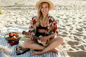 romantico bionda donna nel cannuccia cappello seduta su copertina su il spiaggia e giocando ukulele chitarra. foto