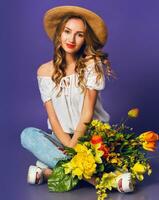 ritratto di bellissimo bionda giovane signora nel elegante cannuccia estate cappello Tenere colorato primavera fiore mazzo vicino viola parete sfondo. foto