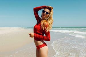 vicino su estate ritratto di sognante contento biondo donna nel rosso bikini in posa su vuoto sorprendente spiaggia vicino oceano. esotico isola. la libertà e vacanza concetto. foto