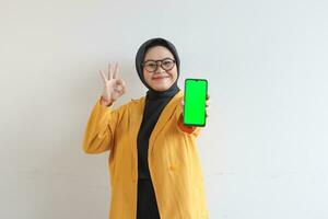 bellissimo giovane asiatico donna nel occhiali, hijab e indossare giallo blazer mostrando verde schermo mobile Telefono mentre sorridente foto