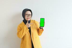 bellissimo giovane asiatico donna nel occhiali, hijab e indossare giallo blazer è Tenere verde schermo mobile Telefono con cupo viso foto