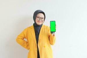 bellissimo giovane asiatico donna nel occhiali, hijab e indossare giallo blazer è Tenere verde schermo mobile Telefono con contento viso foto