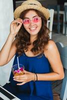 attraente giovane donna nel blu vestito e cannuccia cappello indossare rosa occhiali da sole potabile cocktail foto