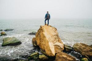 l'uomo viaggiatore con uno zaino si erge su una roccia contro un bellissimo mare foto